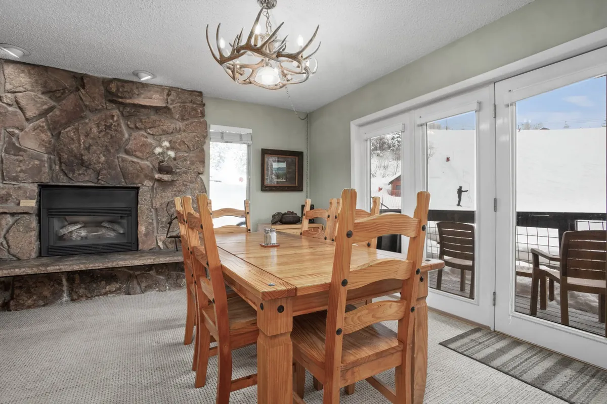 A beautifully set dining table positioned near a fireplace, illuminated by a chandelier. The space is filled with natural light from large windows, hinting at snowy mountain views outside. The room is arranged for group gatherings, game nights, or festive dinners.