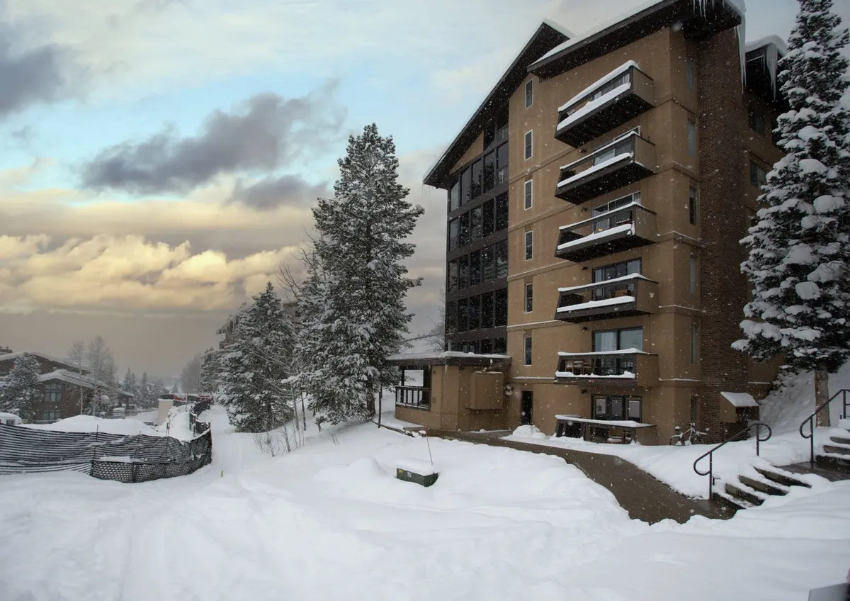 A snow-covered building set among trees, with a snowy pathway leading up to it. The exterior suggests a cozy retreat after an active day, while inviting guests to gather and socialize. The image hints at shared outdoor amenities like fire pits and communal areas.