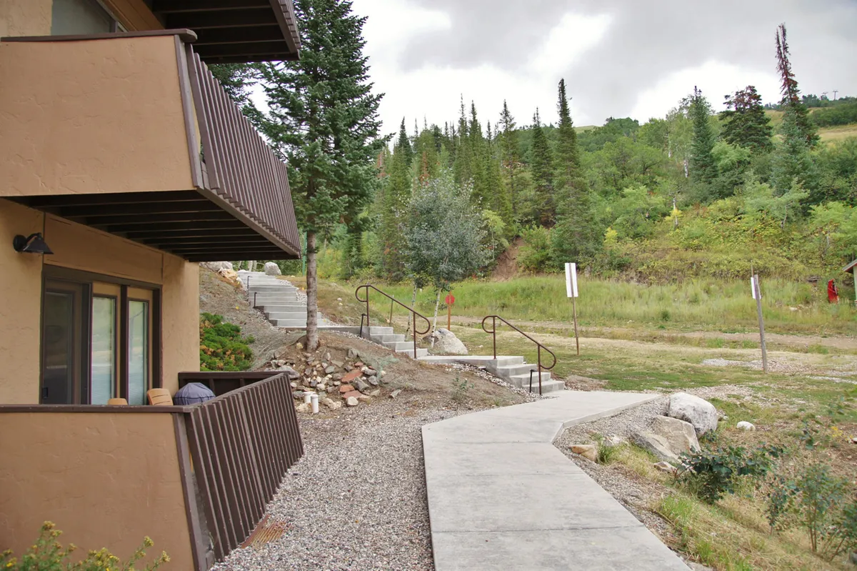 The image showcases the condo’s charming exterior nestled among tall, lush trees. The inviting pathway and staircase lead right up to the front door, highlighting easy access for outdoor adventures. The serene setting promises peaceful moments and breathtaking mountain views, with plenty of space for gatherings on the patio.