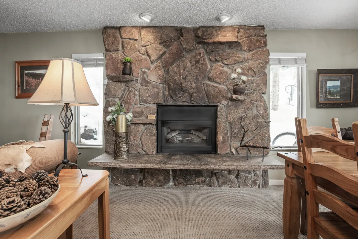 A warmly lit, spacious interior with a prominent stone fireplace, a large wooden dining table set for gatherings, and inviting décor. The room exudes a cozy, communal atmosphere perfect for family meals or sharing stories by the fire. Artistic accents and comfortable seating make it ideal for relaxing after a day on the mountain.