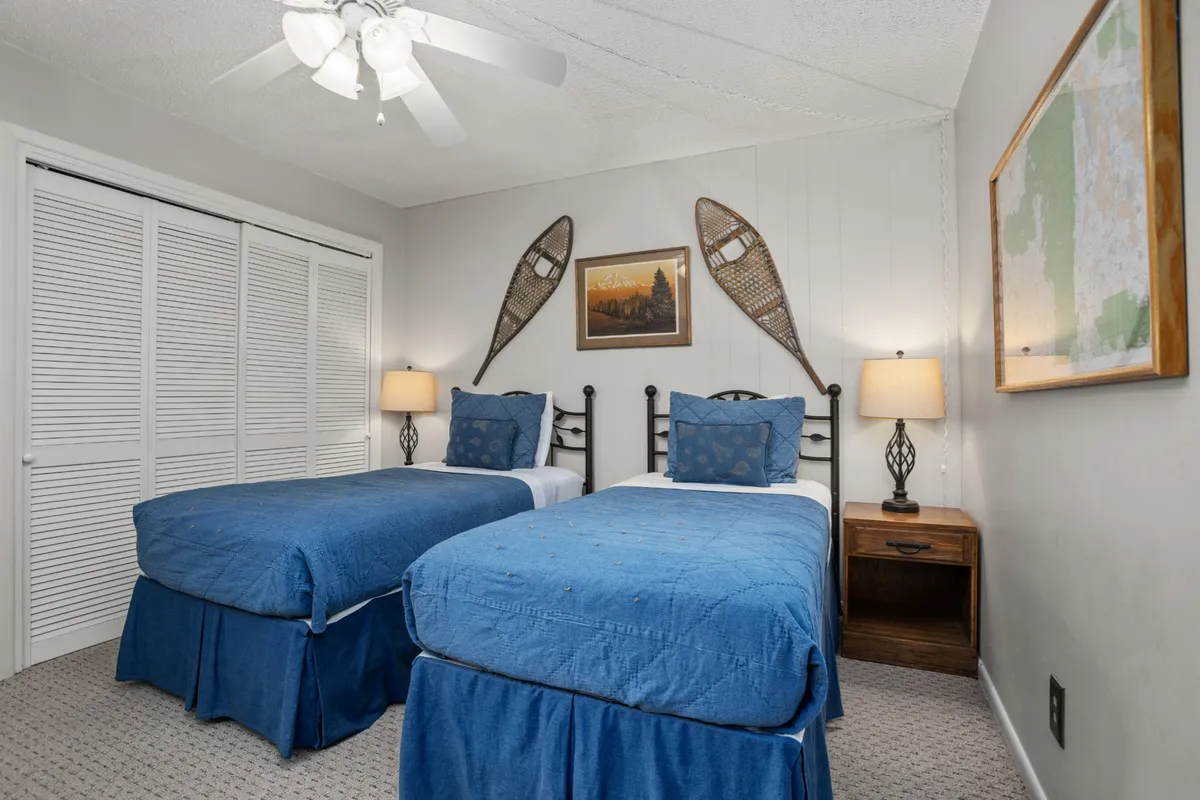 A cozy bedroom with twin beds, snowshoes mounted on the wall, bedside lamps casting a warm glow, and inviting framed artwork. The room is arranged to accommodate multiple sleepers in comfort and style, perfect for flexible group arrangements.