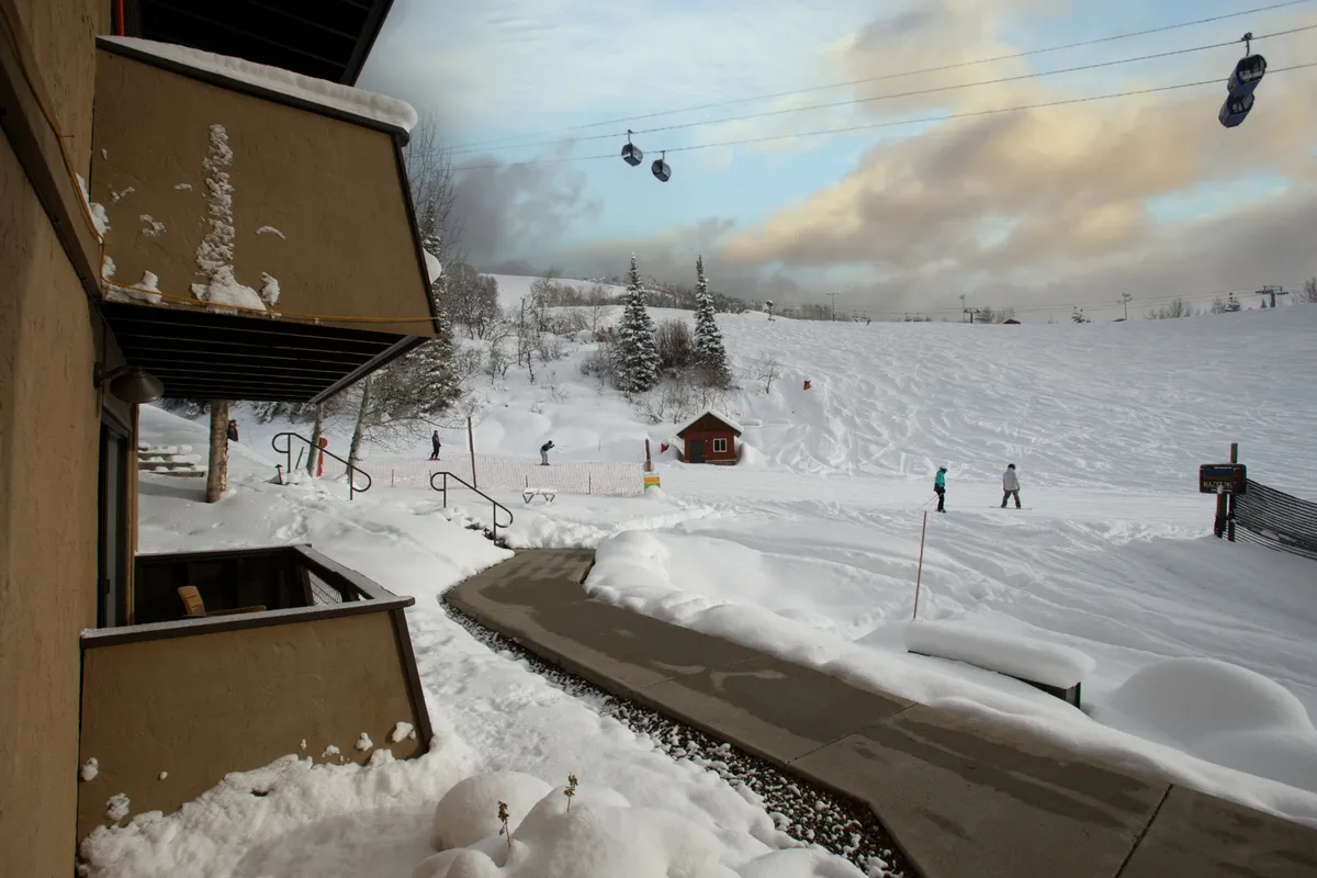 A snow-covered walkway leading to the slopes, with ski lift gondolas overhead, trees dusted in snow, and benches lining the path. The scene captures the heated walkway and the immediate, inviting access from the residence to the ski area.