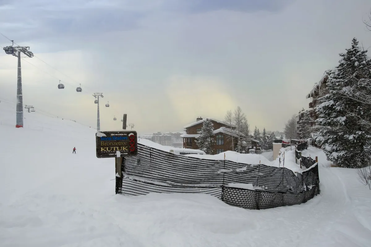 An exterior view highlighting a ski lift ascending a snow-covered slope, flanked by wooden lodge buildings and snow fencing, all under a serene winter sky. The scene invites thoughts of exploration and family outings.