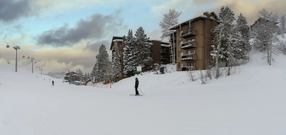 This exterior image highlights ski slopes peppered with skiers and snowboarders, snow-covered trees, and buildings with balconies nestled right at the base. The scene radiates action and accessibility, situating the viewer in the heart of a bustling mountain resort.