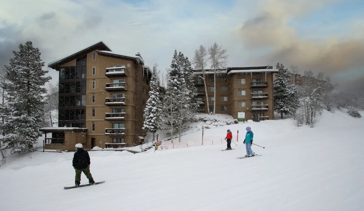 A picturesque snowy landscape featuring skiers gliding down the slopes, the building’s facade visible among trees dusted with fresh snow. The scene blends adventure with coziness, hinting at fun-filled days and relaxing evenings nearby.