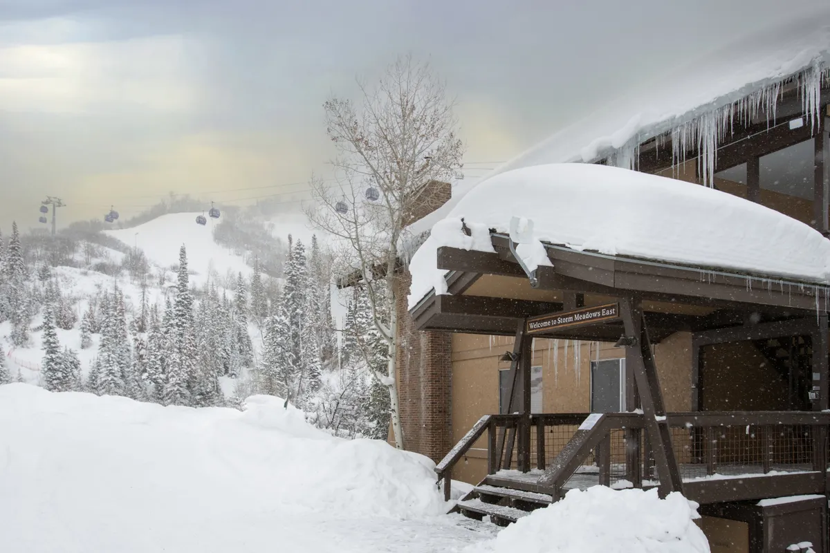 The snow-covered roof and icicle-adorned eaves of a wooden porch frame a tranquil, welcoming entryway. The signage and cozy porch suggest warmth and hospitality just steps from the winter action.