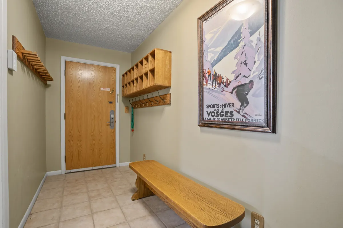 A functional and inviting entryway featuring a wooden bench for changing footwear, wall-mounted coat racks for hanging jackets and ski gear, a vintage ski poster adding a touch of mountain nostalgia, and a sturdy front door. The space is designed to handle snowy boots and bulky outerwear, perfect for seamless transitions after a day on the slopes.