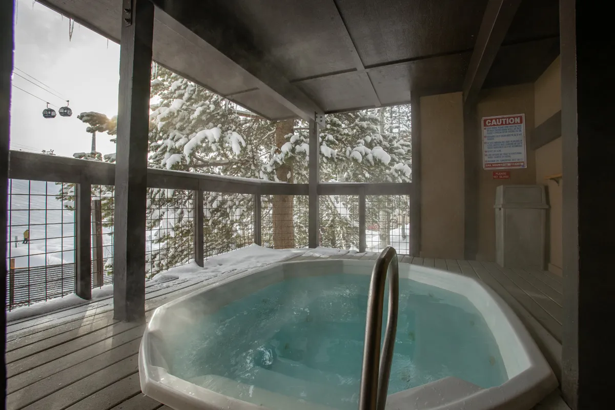 A steaming outdoor hot tub sits amid a picturesque snowy landscape, with cable cars gliding by in the background. The setting is both serene and social, inviting guests to relax and soak after a day on the mountain.