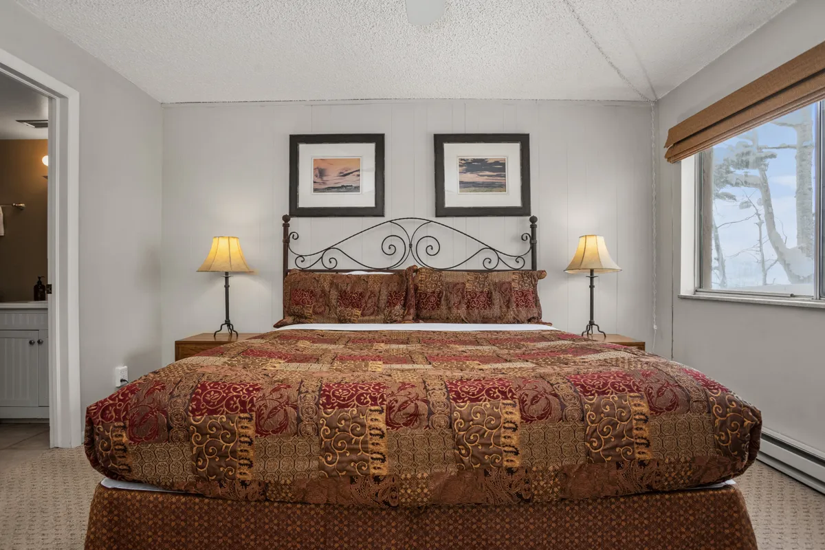 This image shows a neatly made bed with soft linens, flanked by bedside lamps that emit a warm glow. Over the bed, there are calming paintings that add to the tranquil atmosphere. The room basks in natural light, hinting at a restful retreat after a day on the slopes.