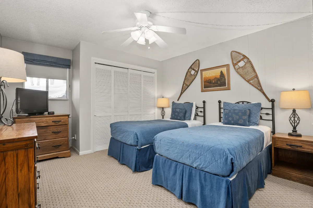 A serene bedroom featuring two beds with calming blue bedding, a wooden dresser, nightstands with lamps, and framed artwork. Snowshoes on the wall add a rustic touch. The room is brightened by natural light, creating a peaceful ambiance ideal for relaxation.