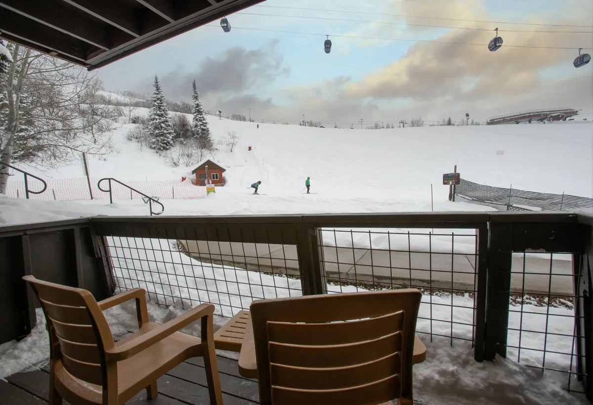 This image shows a private patio with wooden chairs arranged to face the stunning ski slopes in the background. Gondolas glide overhead, snow-covered trees frame the view, and a small red building adds a pop of color. The scene exudes a cozy, adventurous mountain retreat vibe.