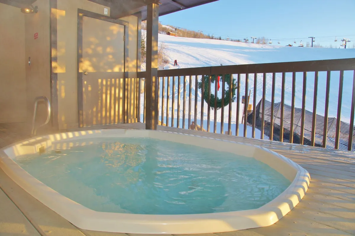 An outdoor hot tub sits invitingly amidst a snowy landscape, with a dramatic view of ski lifts and mountain peaks in the background. The warm steam from the tub contrasts with the snow around it, creating a tranquil, relaxing atmosphere ideal for après-ski relaxation.