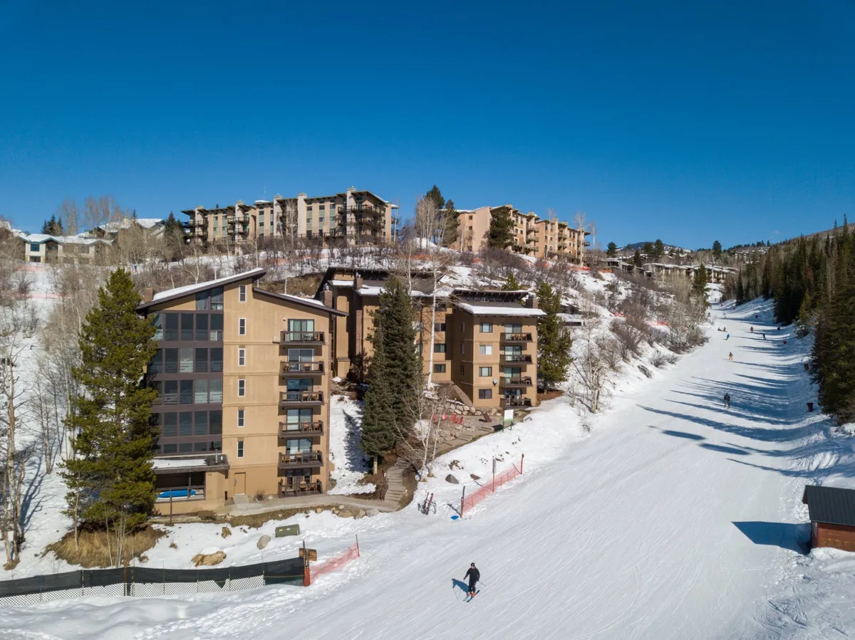 A lively exterior winter scene capturing the ski slopes surrounded by buildings and tall trees, bathed in morning sunlight. People are seen skiing and snowboarding, with the vibrant base area and mountain backdrop conveying both excitement and accessibility.